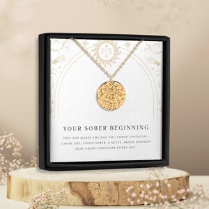 May include: A gold-colored pendant necklace with a circular charm featuring a map design. The necklace is displayed in a black box with the text "YOUR SOBER BEGINNING". The box is on a wooden stand, with pink flowers in the foreground.