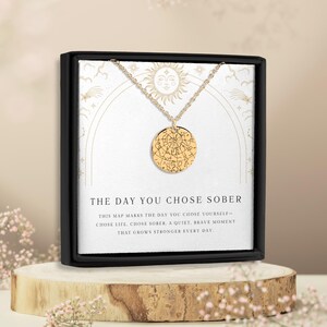 May include: Gold-colored pendant necklace with a circular charm featuring a map design. The necklace is displayed in a black box with the text "THE DAY YOU CHOSE SOBER". The box is on a wooden stand, with floral accents.