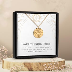 May include: A gold-colored pendant necklace with a circular charm featuring a map design. The necklace is displayed in a black box with the text "YOUR TURNING POINT". The box is on a wooden stand, with floral accents.