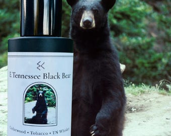 May include: A cylindrical glass bottle of E Tennessee Black Bear Eau de Parfum with a black cap. The white label features a black bear illustration, the text "E Tennessee Black Bear", "Cedarwood • Tobacco • TN Whiskey", and "1.7 fl oz / 50 mL".