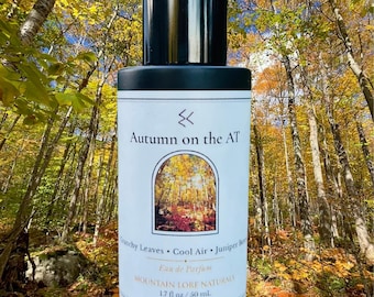 Peut inclure: Un flacon de parfum cylindrique avec un bouchon noir et une étiquette blanche. L'étiquette porte le texte Autumn on the AT, Mountain Lore Naturals, 1.7 fl oz / 50 ml, et une petite illustration arquée d'une forêt. Le texte Crunchy Leaves, Cool Air, Juniper Berry est également visible.