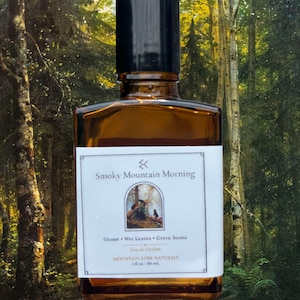 May include: A rectangular amber glass perfume bottle with a black cap and a white label. The label reads Smoky Mountain Morning, Ozone, Wet Leaves, Green Stems, Eau de Parfum, Mountain Lore Naturals, and 1 fl oz / 30 mL with a small forest graphic.