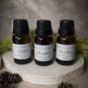May include: Three small, dark glass bottles with black caps and white labels, each labeled with a different botanical name: "Fern," "Meadow," and "Flora." The bottles are displayed on a textured, white surface, with pine cones scattered around.