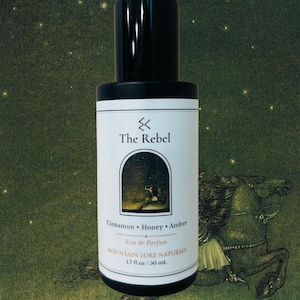 May include: A black and white perfume bottle labeled "The Rebel" with the scent notes of Cinnamon, Honey, and Amber. The bottle is 1.7 fl oz / 50 ml. The label features an illustration of a person in an archway. The background is a painting of a horse.