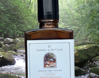 May include: A rectangular amber glass perfume bottle with a black cap and a white label. The label features the text Summer at the Creek, Humid Air, Rain, Fresh Soil, Eau de Parfum, MOUNTAIN LORE NATURALS, and 1 fl oz / 30 ml with a small image.