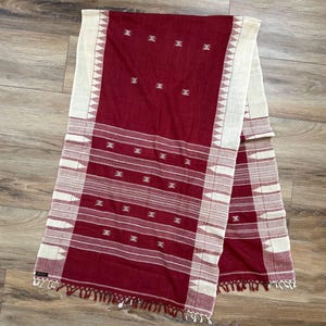 May include: A rectangular woven textile featuring a deep maroon center panel with small white geometric motifs and horizontal stripes. The edges are bordered by wide cream-colored bands with triangular patterns and fringed ends at the bottom.