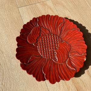 May include: A round, red, carved wooden decorative plate in the shape of a flower. The plate has detailed petal and center designs. The plate is on a light brown wooden surface.
