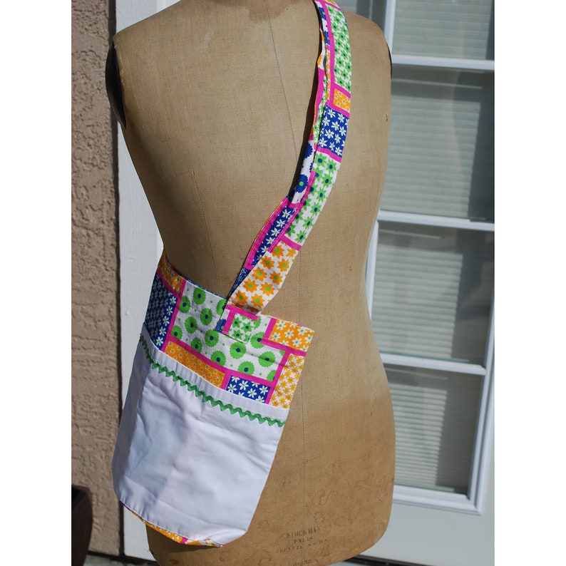 Puede incluir: Una bolsa de tela blanca con un dise&ntilde;o de patchwork en tonos de rosa, verde, azul y amarillo. La bolsa tiene una correa larga y un forro blanco.