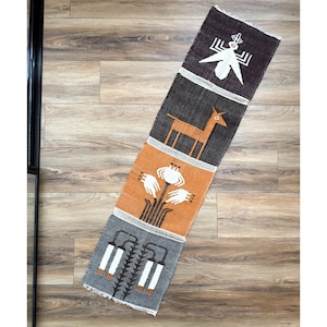 May include: A long, rectangular woven table runner featuring four distinct horizontal panels. The designs include a white insect, a tan deer, a white floral motif on an orange background, and white hanging lanterns on a gray background.