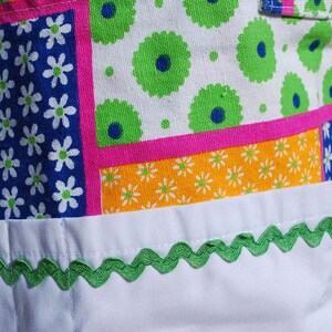 Puede incluir: Una bolsa de tela blanca con un dise&ntilde;o de patchwork que presenta varios patrones florales en tonos azules, verdes, rosas y amarillos. La bolsa tiene un ribete festoneado verde alrededor del borde superior.