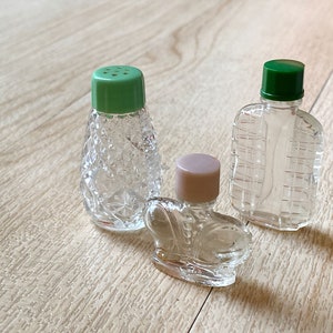 May include: Three small glass bottles with different designs and colored lids. The bottles are clear and have a textured surface. The lids are green and pink.