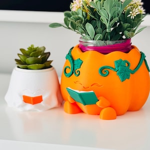 May include: A white ghost-shaped planter containing a green succulent and an orange pumpkin-shaped planter with green vine details containing a green plant. Both planters feature a small character reading a book, with the ghost holding an orange book.