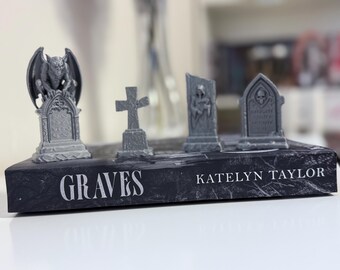 May include: A book titled "Graves" by Katelyn Taylor, with miniature gray tombstones and a gargoyle statue on top. The tombstones include a cross, a broken tombstone with a skeleton, and a tombstone with a skull.