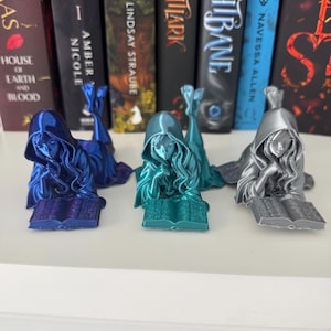 May include: Three resin bookends in blue, teal, and silver. Each depicts a hooded figure reclining and reading a book. The bookends are displayed in front of a row of books. The bookends are made of resin.