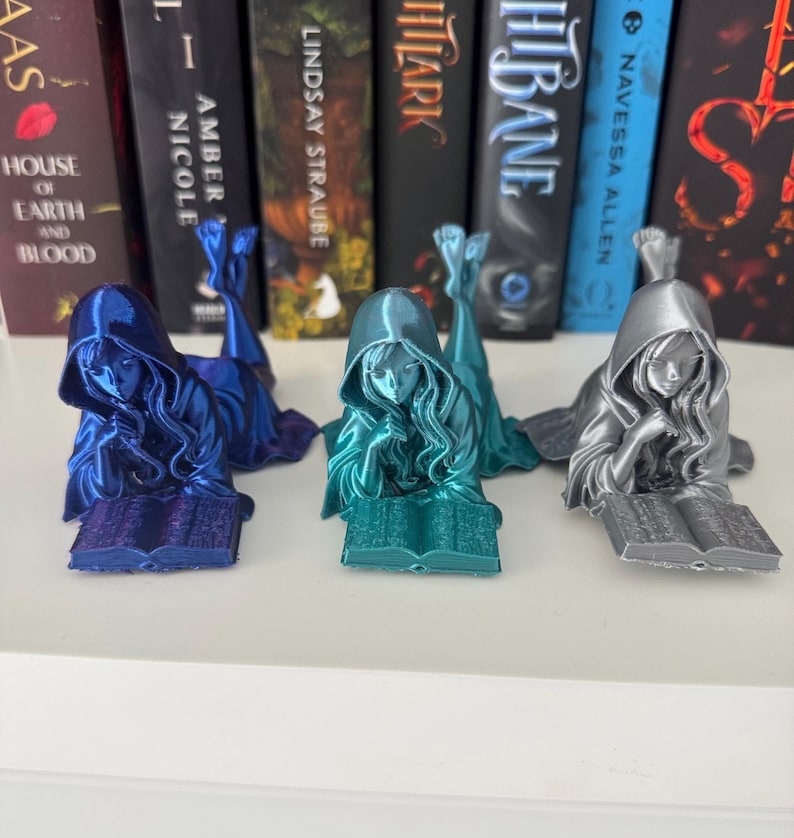 May include: Three resin bookends in blue, teal, and silver. Each depicts a hooded figure reclining and reading a book. The bookends are displayed in front of a row of books. The bookends are made of resin.