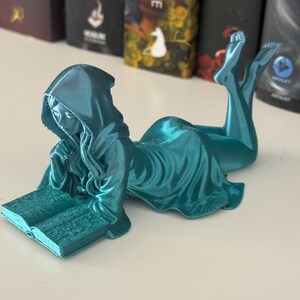 May include: A teal figurine of a hooded figure reading a book while reclining. The figure is wearing a flowing robe and is posed with legs extended. The book is open, and the figure appears to be engrossed in reading. The figurine is on a white surface.
