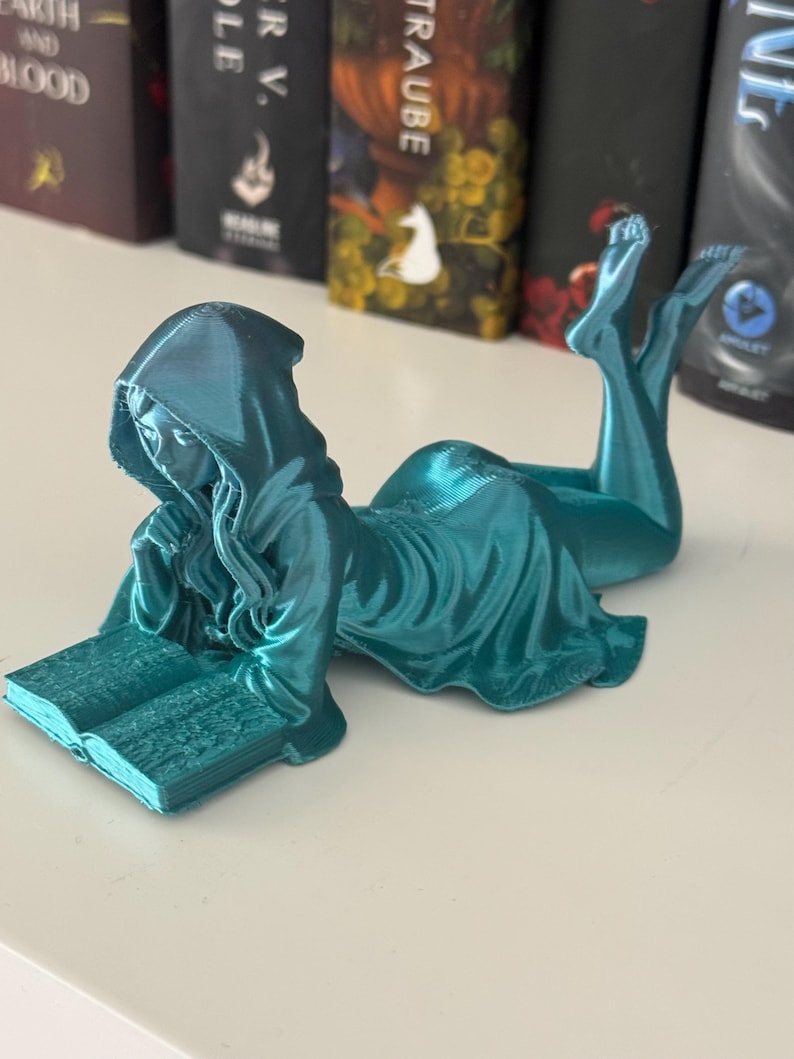 May include: A teal figurine of a hooded figure reading a book while reclining. The figure is wearing a flowing robe and is posed with legs extended. The book is open, and the figure appears to be engrossed in reading. The figurine is on a white surface.