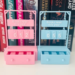 May include: Two book counter organisers, one pink and one blue, each with a drawer and a counter that reads "Books read this year." The pink counter shows "012," and the blue counter shows "055." The organisers are set against a backdrop of colourful books.