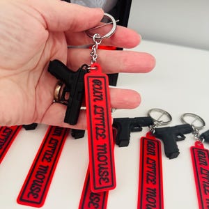 May include: Black gun-shaped keychains with red tags that read "Run Little Mouse" in white lettering. The keychains are attached to silver keyrings. The image shows multiple keychains, with one held in a hand.