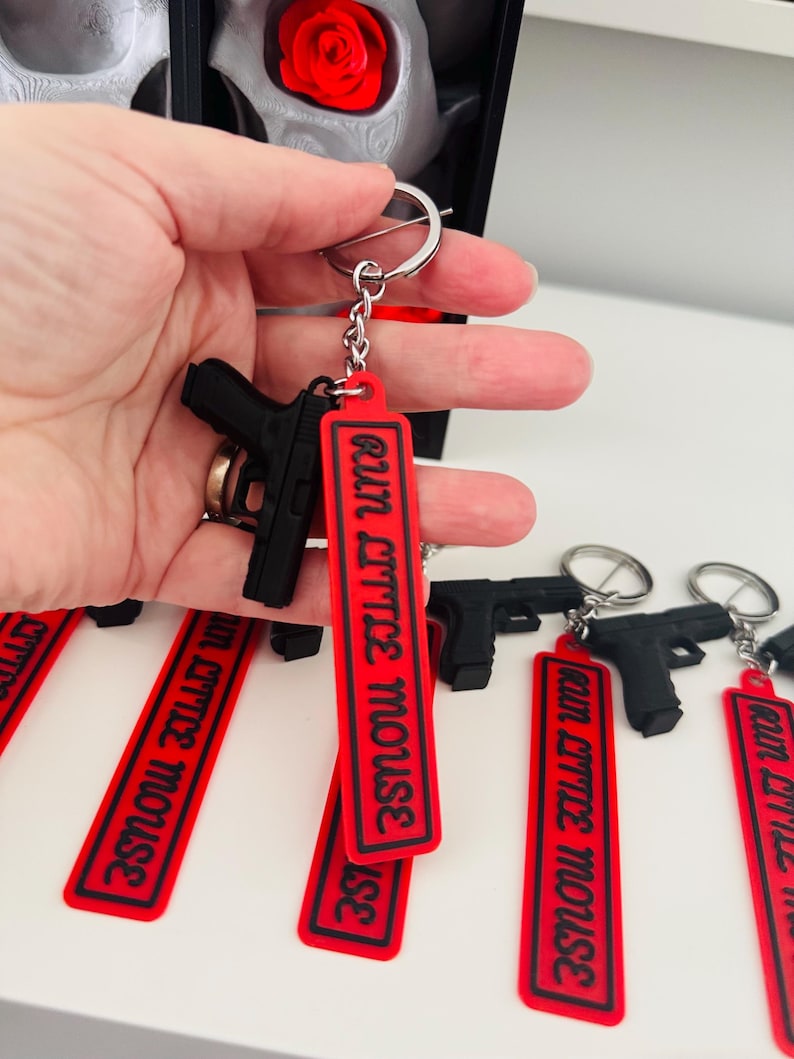 May include: Black gun-shaped keychains with red tags that read "Run Little Mouse" in white lettering. The keychains are attached to silver keyrings. The image shows multiple keychains, with one held in a hand.