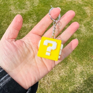 May include: A yellow keychain with a white question mark on a square block. The keychain has a silver ring and chain. The background is a blurred view of green grass.
