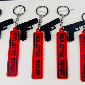 May include: Black gun-shaped keychains with red rectangular tags. The tags have the phrase "RUN LITTLE MOUSE" in black lettering. Each keychain has a silver-colored ring and chain.