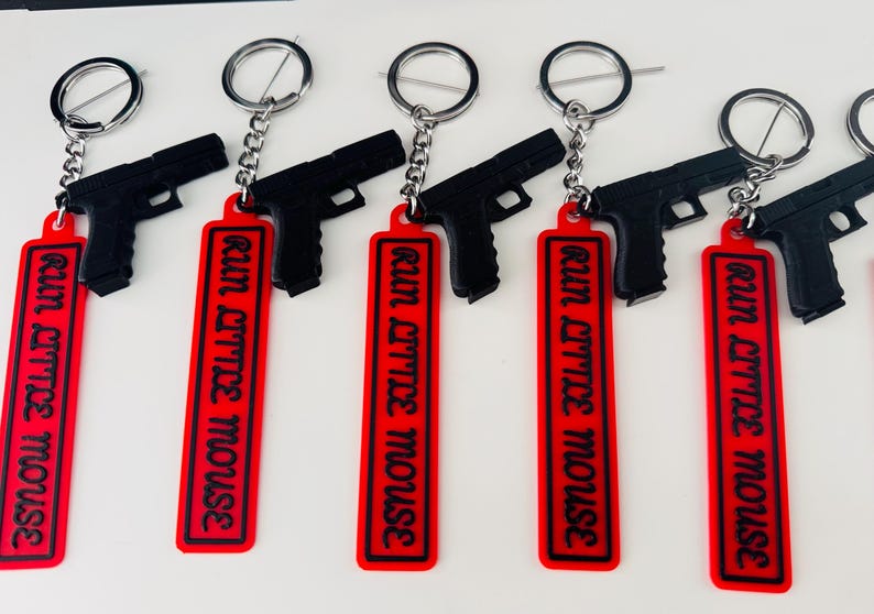 May include: Black gun-shaped keychains with red rectangular tags. The tags have the phrase "RUN LITTLE MOUSE" in black lettering. Each keychain has a silver-colored ring and chain.