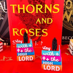 May include: A collection of six miniature book ornaments featuring stacks of books with text. Two ornaments feature black top hats and the text "IT'S ONLY A GAME". Two feature black figures and the text "stay with the HIGH LORD". Two feature black dragons and the text "FLY OR DIE".