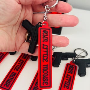 May include: Black handgun keychain with a red tag that reads "run little mouse" in white text. The keychain is attached to a silver keyring. Several similar keychains are visible.