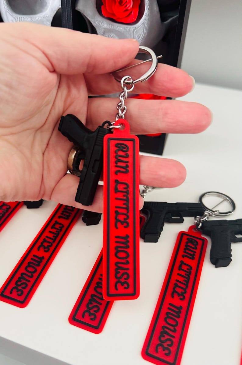 May include: Black handgun keychain with a red tag that reads "run little mouse" in white text. The keychain is attached to a silver keyring. Several similar keychains are visible.