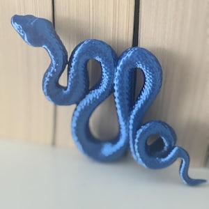 May include: A metallic blue snake figurine with a textured, scaled surface. The snake is depicted in a winding, S-shaped posture with its head raised and pointed towards the left, showcasing a glossy finish and detailed, realistic body contours.