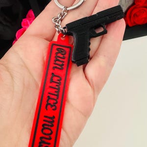 May include: Black handgun keychain with a red rectangular tag. The tag has the phrase "run little mouse" in black lettering. The keychain is attached to a silver metal ring.