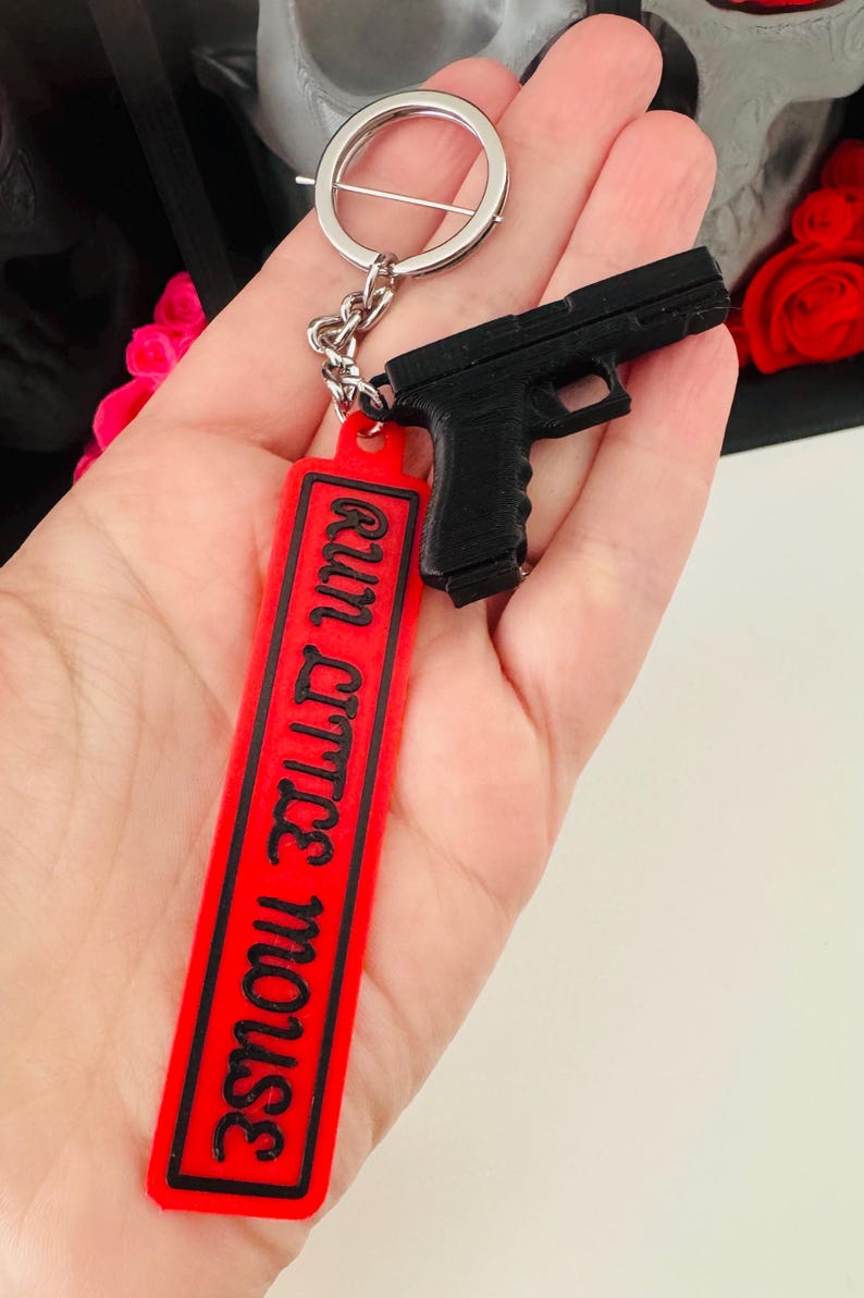 May include: Black handgun keychain with a red rectangular tag. The tag has the phrase "run little mouse" in black lettering. The keychain is attached to a silver metal ring.
