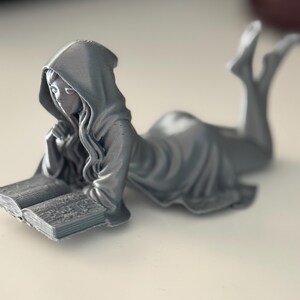 May include: A grey figurine of a person in a hooded robe, reclining and reading a book. The figure is lying down, with legs raised, and is holding the book with both hands. The book is open, and the pages are detailed. The figurine is on a white surface.