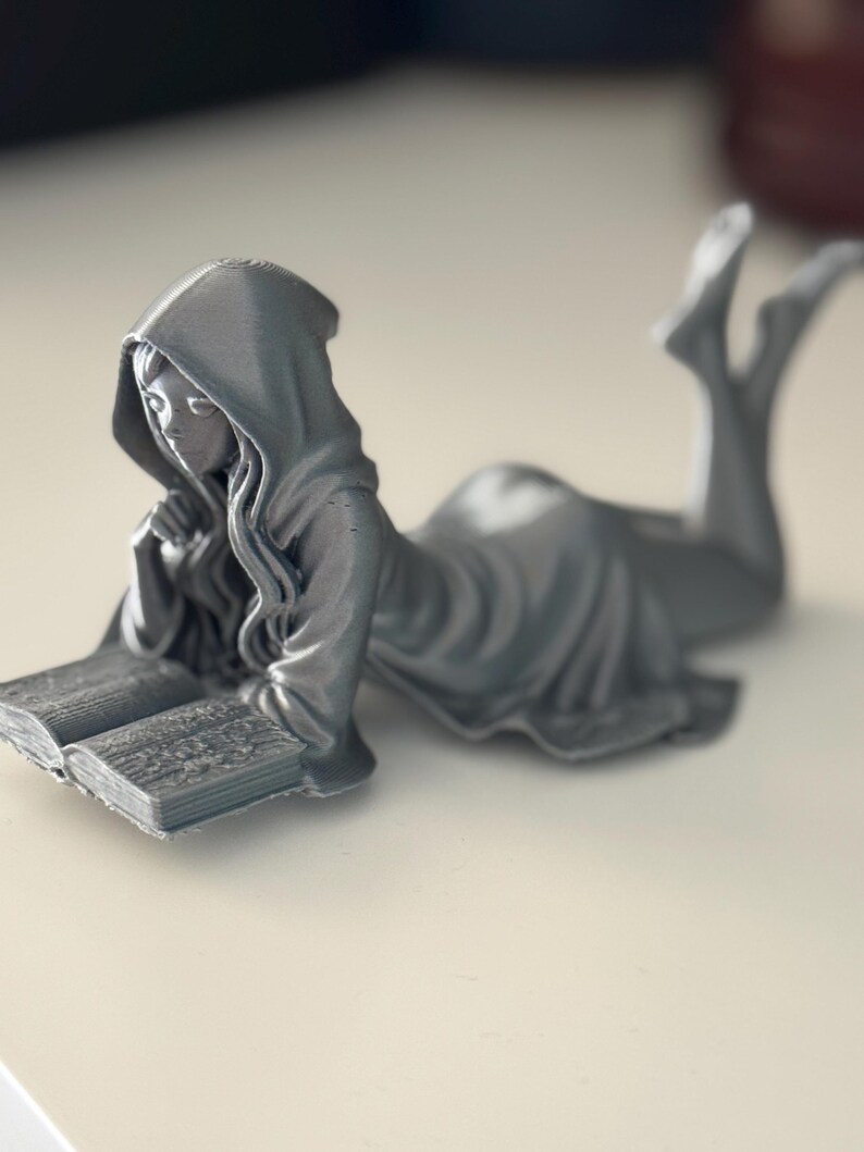 May include: A grey figurine of a person in a hooded robe, reclining and reading a book. The figure is lying down, with legs raised, and is holding the book with both hands. The book is open, and the pages are detailed. The figurine is on a white surface.