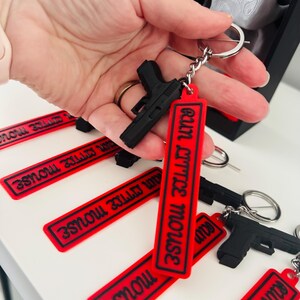 May include: Black gun-shaped keychains with red rectangular tags that read "EATIN LITTLE MOUSE" in black lettering. The keychains are attached to silver keyrings. Several keychains are visible, some held in a hand.