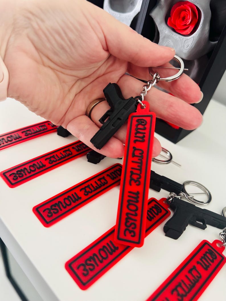 May include: Black gun-shaped keychains with red rectangular tags that read "EATIN LITTLE MOUSE" in black lettering. The keychains are attached to silver keyrings. Several keychains are visible, some held in a hand.