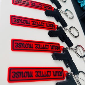May include: Black pistol-shaped keychains with silver key rings attached to red rectangular tags. The tags have the text "RUN MITTIE MOUSE" in black. The keychains are displayed against a white background.