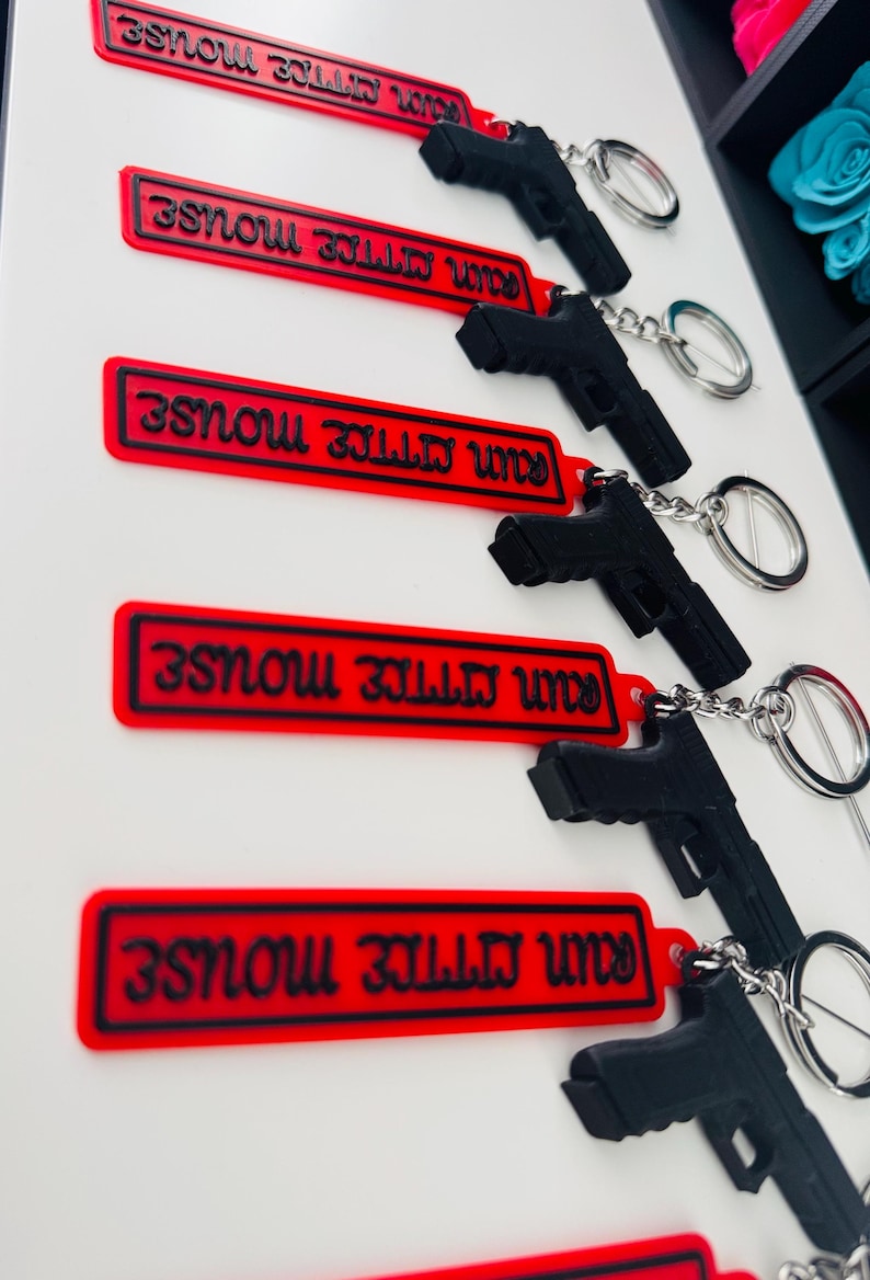 May include: Black pistol-shaped keychains with silver key rings attached to red rectangular tags. The tags have the text "RUN MITTIE MOUSE" in black. The keychains are displayed against a white background.