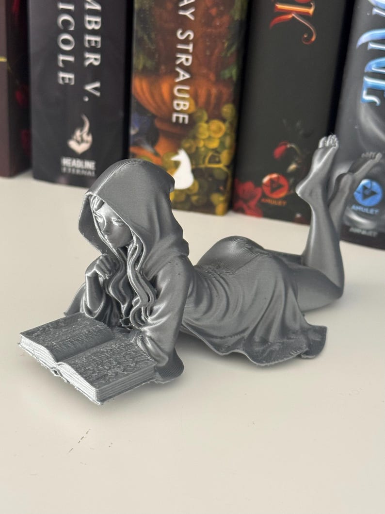 May include: A silver figurine of a person in a hooded robe, reading a book while reclining. The figure has flowing robes and long hair. The open book has visible pages. Books are in the background, creating a literary theme.