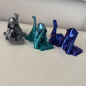 May include: Three metallic figurines of a hooded figure reclining and reading a book. The figurines are in silver, teal, and blue. The figures are lying down with their legs in the air, and the book is open in front of them.