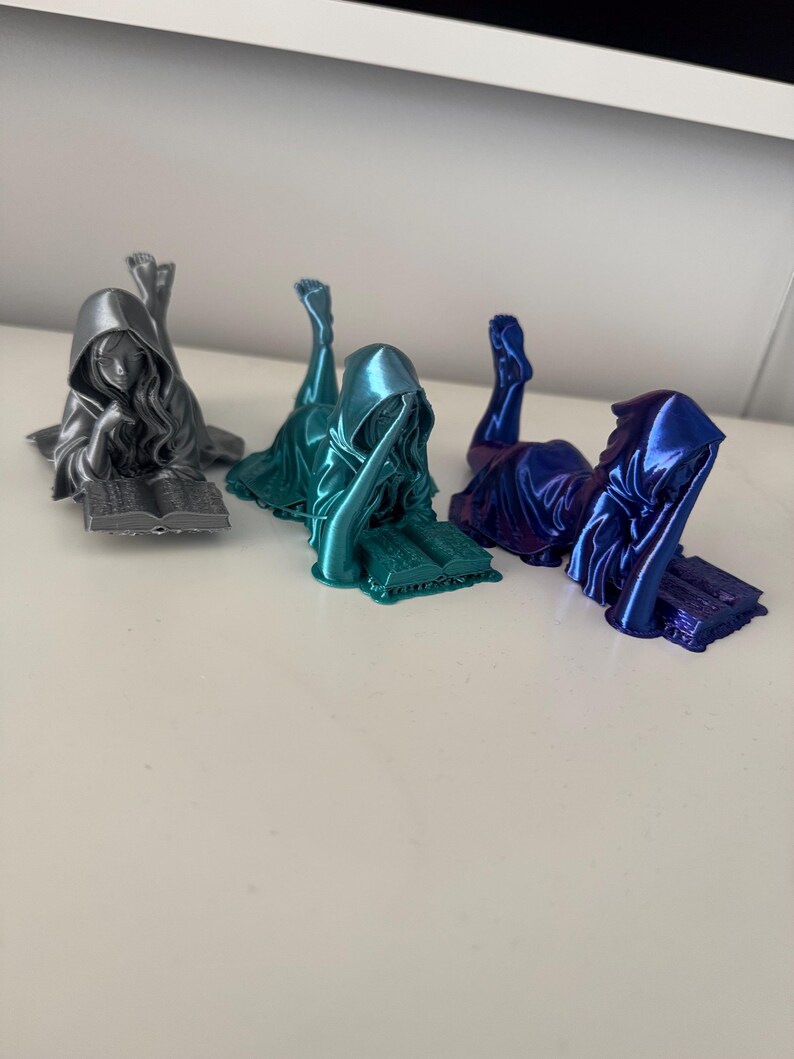 May include: Three metallic figurines of a hooded figure reclining and reading a book. The figurines are in silver, teal, and blue. The figures are lying down with their legs in the air, and the book is open in front of them.