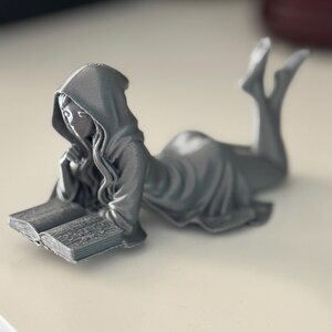 May include: A silver figurine depicting a person in a hooded robe, reading a book while reclining. The figure is lying on their front, with legs extended and feet pointing upwards. The open book is in front of them, with their chin resting on their hand.