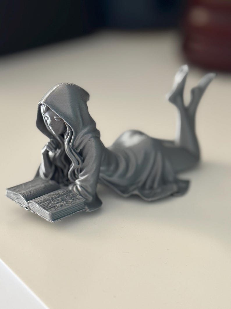 May include: A silver figurine depicting a person in a hooded robe, reading a book while reclining. The figure is lying on their front, with legs extended and feet pointing upwards. The open book is in front of them, with their chin resting on their hand.