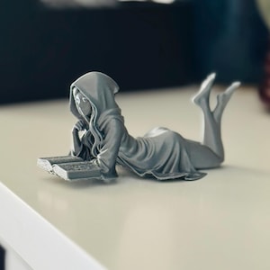 May include: A grey figurine of a person in a hooded robe, reclining and reading a book. The figure is lying on its stomach with its legs raised. The book is open, and the figure's face is visible. The figurine is on a white surface.