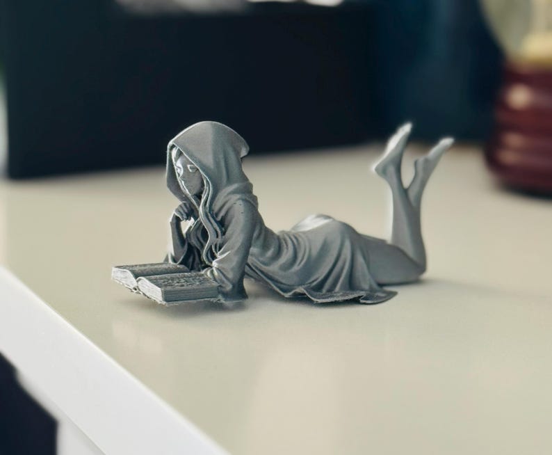 May include: A grey figurine of a person in a hooded robe, reclining and reading a book. The figure is lying on its stomach with its legs raised. The book is open, and the figure's face is visible. The figurine is on a white surface.