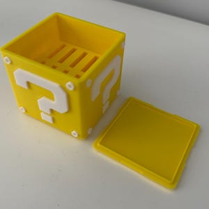 May include: A yellow plastic question mark block with a white question mark on each side. The block is open and the lid is off. The block has a small compartment inside.