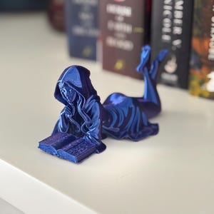 May include: A blue figurine of a hooded figure reclining and reading a book. The figure is on a white surface, with the spine of several books in the background. The figurine has a metallic sheen.