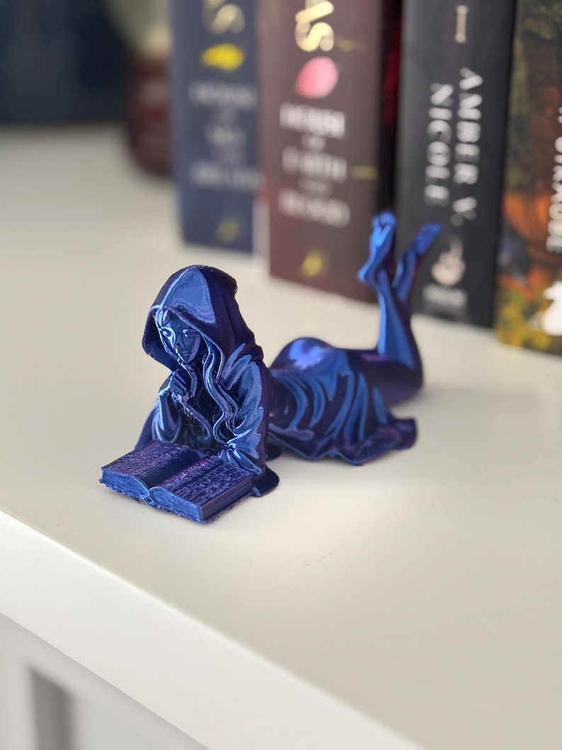 May include: A blue figurine of a hooded figure reclining and reading a book. The figure is on a white surface, with the spine of several books in the background. The figurine has a metallic sheen.