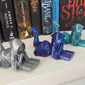 May include: Three figures of a hooded person reading a book. The figures are in silver, blue, and teal. The figures are lying down, with their legs extended. Books are in the background.
